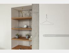 Eingelassene Holzregale in einer weißen Wandnische mit Betonrückwand, dekoriert mit Pflanzen, einer Uhr und kleinen Objekten; daneben eine weiße Wand mit einem aufgehängten Drahtkleiderbügel.