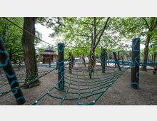Kletterspielplatz im Freien mit Holzpfosten, die durch Seilnetze verbunden sind, umgeben von Bäumen und Laub. Im Hintergrund ist ein hölzerner Pavillon sichtbar.