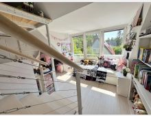 Helles Dachgeschoss-Kinderzimmer mit weißem Holzfußboden, großem Fensterband und Tagesbett unter den Fenstern. Im Vordergrund führt eine offene Treppe mit Holzhandlauf und Seilgeländer in den Raum; rechts stehen Regale mit Büchern und Dekoration.