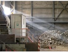 Eine Industriehalle mit einer kleinen, grauen Kabine, ausgestattet mit einem Erste-Hilfe-Symbol und einer Außentreppe mit rotem Geländer. Im Hintergrund sind ein großer Schutthaufen und Metallträger an der Betonwand sichtbar, während Tageslicht durch eine Öffnung einfällt.