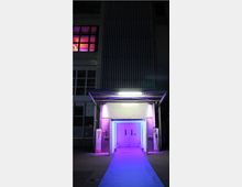 Industriegebäude bei Nacht mit beleuchtetem Eingang und blauem Teppich. Der Eingang ist von lila und blauen Lichtakzenten umgeben, während ein großes Fenster im oberen Bereich des Gebäudes ebenfalls in lila Licht leuchtet.