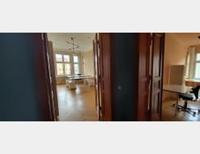 Blick durch einen Flur in ein helles Büro mit Fischgrät-Holzboden, hohen Decken und großen Sprossenfenstern. Im Raum stehen mehrere Schreibtische und Bürostühle; rechts sind geöffnete dunkle Holztüren und ein weiterer Arbeitsbereich zu sehen.