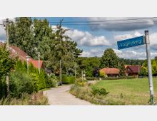 Eine ländliche Wohngegend mit einer schmalen Straße, gesäumt von Bäumen und Sträuchern sowie Häusern mit roten Dächern. Im Vordergrund ein Straßenschild mit der Aufschrift „Vogelweg“, im Hintergrund Wiesen und dichte Vegetation.