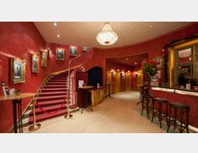 Eingangsbereich eines Theaters oder Varietés mit roten Wänden, einer geschwungenen Treppe mit rotem Teppich und goldenen Geländern. Der Raum ist dekoriert mit vergoldeten Bilderrahmen, einem Kronleuchter und einer Bar auf der rechten Seite.