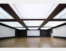 Rechteckiger, leerer Raum mit dunklen Wänden und hellem Holzfußboden, beleuchtet durch große, rechteckige Deckenleuchten mit einem Rastermuster. Eine Tür befindet sich an der rechten Wand, und die Rückwand ist durch rechteckige, weiße Flächen akzentuiert.