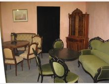 Ein Raum mit antikem Mobiliar, darunter ein grünes Sofa, mehrere gepolsterte Stühle, ein runder Holztisch und ein Vitrinenschrank aus Holz. Im Hintergrund eine schwarze Türöffnung und eine Wand in hellrosa Farbe mit einem gerahmten Bild.