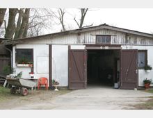 Ein einfaches landwirtschaftliches Nebengebäude mit weißer Fassade und braunen Holztüren, von denen eine offen steht und einen Blick ins Innere ermöglicht. Vor dem Gebäude stehen eine Schubkarre, vereinzelte Stühle sowie Blumenkästen mit Tannenzweigen als Dekoration.