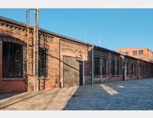 Long red-brick industrial building with arched, metal-barred windows and a large gray door, bordering a cobblestone courtyard. A vertical metal ladder and rooftop gutters line the facade under a clear blue sky, with another brick building visible in the background.