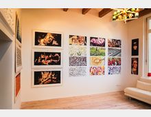 Ein heller Ausstellungsraum mit Holzboden, weißen Wänden und einer weißen Couch. An den Wänden hängen mehrere bunte Gemälde und Zeichnungen, die verschiedene Motive wie Feuer, Menschen, Blumen und abstrakte Muster zeigen.