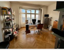 Ein Arbeitszimmer mit einem großen Schreibtisch, mehreren Monitoren und einem Bürostuhl auf einem Holzboden. Im Raum befinden sich ein Regal mit Büromaterialien, ein Aktenschrank und hohe Fenster, durch die Tageslicht einfällt und eine Aussicht auf die Stadt sichtbar ist.