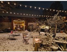 Schneebedeckter Hof mit Lichterketten, Laternen und winterlicher Dekoration; im Hintergrund ein geöffneter Scheuneneingang mit weiterer Beleuchtung, umgeben von Bäumen und Pflanzen. Tische und Stühle stehen verteilt im Außenbereich.