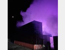 Zwei übereinandergestapelte Frachtcontainer bei Nacht, umgeben von lila, beleuchtetem Rauch, mit einem beleuchteten Mond im Hintergrund. Im Vordergrund ist ein Dreibein-Stativ erkennbar.