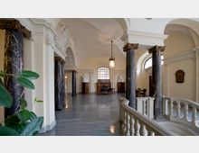 Innenansicht eines historischen Gebäudes mit hohen, gewölbten Decken, großen dunklen Marmorsäulen und dekorativen Stuckelementen. Im Hintergrund steht eine Sitzgruppe vor einem großen Fenster sowie weißen Büsten auf Holzsockeln, während im Vordergrund ein Balustradengeländer sichtbar ist.