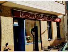 Straßenansicht eines kleinen Geschäfts mit dem Schild "La Bohème artistique" über dem Eingang. Das Gebäude hat ockerfarbene Wände, blaue Fensterrahmen und eine große Schaufensterscheibe mit Pflanzen im Inneren; im Vordergrund steht ein alter Wasserhydrant.