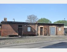 Einstöckiges Backsteingebäude mit flachem Dach, mehreren Türen aus Holz und Metall sowie Fenstern. Der Boden davor ist mit Kopfsteinpflaster und Schienen bedeckt, im Hintergrund sind Bäume erkennbar.