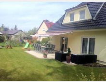 Ein Einfamilienhaus mit heller Fassade und dunklem Satteldach, umgeben von einem Garten mit Rasenfläche. Eine Terrasse mit Sitzmöbeln, einem langen Esstisch und einer Markise befindet sich direkt vor dem Haus; im Hintergrund sind weitere Häuser und ein kleiner Spielplatz mit einer Rutsche zu sehen.