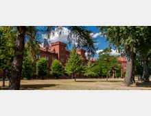 Rotes Backsteingebäude im Stil der Neogotik mit Türmen und vielen Fenstern, umgeben von Rasenflächen und großen Bäumen; blauer Himmel mit weißen Wolken im Hintergrund.