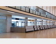 Offener Bereich eines Flughafenterminals mit einer Reihe von verlassenen Check-in-Schaltern, dahinter verglaste Wände und minimalistische Architektur. Über den Schaltern befinden sich Schilder mit Logos verschiedener Fluggesellschaften, und die Etage darüber ist durch eine gläserne Brüstung abgegrenzt.