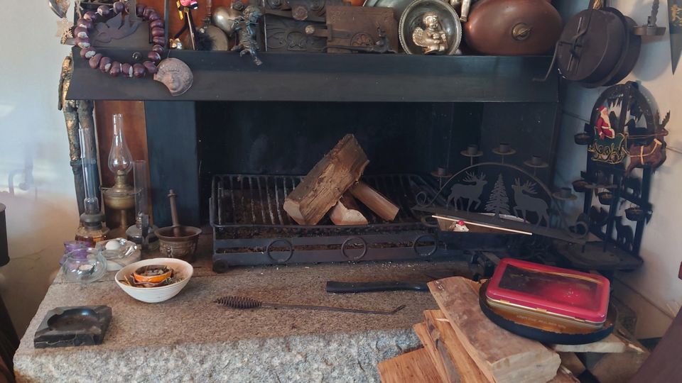 Ein gemütlicher Innenraum mit einer offenen Feuerstelle, umrahmt von einem Holzrahmen und einer Steinplatte. Auf der Feuerstelle stehen und hängen zahlreiche Dekorationsgegenstände, wie Figuren, Metallplatten und Keramiken, während darunter aufgestapeltes Brennholz und ein Korb mit Holzscheiten zu sehen sind.