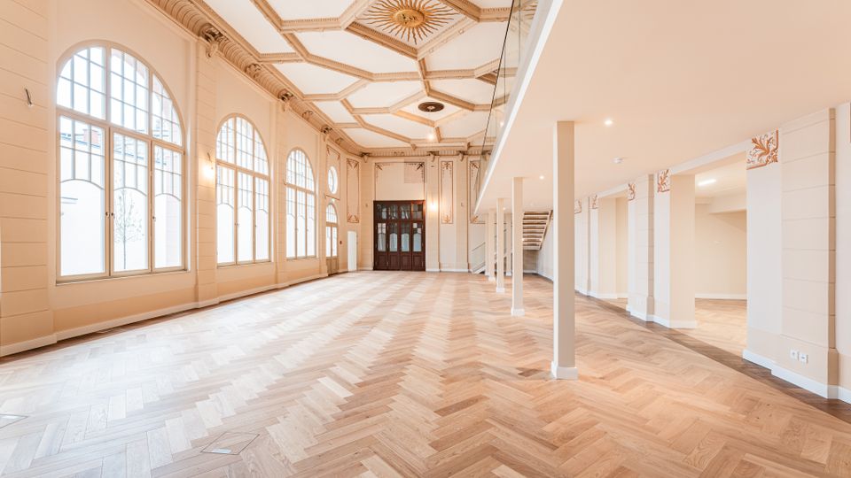 Heller, hoher Saal mit großen, bogenförmigen Fenstern, Fischgrätparkett und verzierter Kassettendecke mit zentralem Ornament. Rechts befindet sich eine offene Galerieebene mit Glasbrüstung, Säulen und einer Treppe; am Ende des Raums stehen dunkle Holztüren.