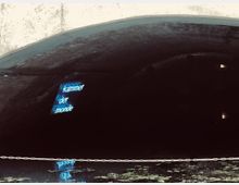 Blick in einen dunklen Tunnel mit einer gewölbten Decke. Im Zentrum hängt ein beleuchtetes Schild mit der Aufschrift „Kammer der Monde“, das sich im Wasser darunter spiegelt; eine Kette ist über das Wasser gespannt.