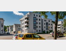 Moderne Wohnanlage mit hellen, mehrstöckigen Gebäuden, Balkonen und schwarzen Fensterrahmen an einer Straßenecke. Im Vordergrund sind parkende Autos und ein Baum, während der Himmel klar und sonnig ist.