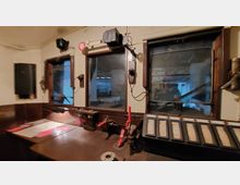 Innenansicht eines historischen Zugführerkabine mit Bedienelementen. Zu sehen sind mehrere Hebel, Schalter, ein Bedienpult mit Beschriftungen und Fenster mit Blick auf den Innenraum einer Werkhalle.