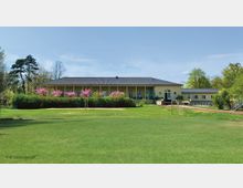 Ein einstöckiges Gebäude mit gelber Fassade und grauem Dach inmitten einer grünen Parklandschaft. Im Vordergrund ein weitläufiger Rasen, umgeben von blühenden Bäumen und Sträuchern, sowie ein schmaler Weg und eine kleine Torstruktur.