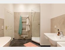 Modernes Badezimmer mit beigen Wänden und dunklem Boden. Sichtbar sind eine freistehende Badewanne, ein Handtuchheizkörper mit aufgehängten Handtüchern, eine verglaste Dusche und ein Waschbecken mit Seifenspender.