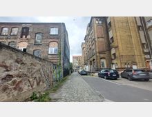Schmale Kopfsteinpflasterstraße zwischen historischen Backsteingebäuden, links verputzte Mauern mit Graffiti, rechts geparkte Autos. Im Hintergrund ist ein weiteres Gebäude mit großen Fenstern sichtbar.