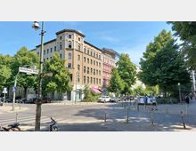 Straßenecke in einer städtischen Umgebung mit mehrstöckigen Wohngebäuden, teils in Pastellfarben, und Bäumen im Vordergrund. Straßenschilder zeigen den Kreuzungspunkt von "Reichenberger Straße" und "Forster Straße"; Fahrräder und parkende Autos sind sichtbar.