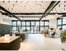 Heller, offener Büroraum mit großen Glasfenstern und Glastüren zur Außenseite, weißen Wänden und einem hellen Boden. Auffällig sind die geometrisch strukturierte Decke mit schwarzen Trägern, eine zentrale Kücheninsel sowie mehrere Arbeitsplätze mit Monitoren und Zimmerpflanzen.