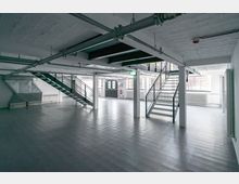 Leerer Industrie-Innenraum mit freiliegenden Metallrohren an der Decke, einer grauen Bodenfläche, zwei metallenen Treppen mit Gittern, weißen Wänden und großen Fenstern, die Tageslicht hereinlassen.