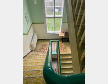 Treppenhaus mit gemustertem Teppichboden, grünem Geländer und großem Fenster, das den Blick auf einen Garten mit Gras, Bäumen und blauen Tonnen freigibt. Ein Holztisch mit einer kleinen braunen Kiste steht am Fenster neben einer Heizkörperverkleidung.