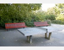 Tischtennisplatte aus Beton mit Metallnetz auf einem gepflasterten Platz, umgeben von zwei roten Holzbänken und dichter Vegetation mit Bäumen und Sträuchern im Hintergrund.