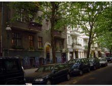 Wohnstraße mit Altbauten in urbaner Umgebung, flankiert von grauen und weißen Gebäuden mit dekorativen Balkonen. Im Vordergrund parken Autos entlang der Straße, während Bäume den Gehweg säumen und Schatten spenden.