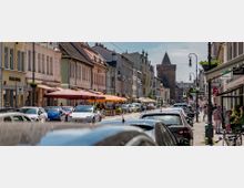 Belebte Altstadtstraße mit historischen Gebäuden, Geschäften und Cafés mit Sonnenschirmen; parkende Autos säumen die Straße, und im Hintergrund ist ein mittelalterlicher Turm sichtbar.