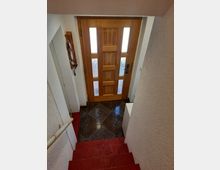 Blick auf einen kleinen Eingangsbereich mit einer Holztür, die seitliche Glaseinsätze hat. Der Boden ist mit dunklen Fliesen ausgelegt, und eine rote Treppe führt hinunter in den Bereich. Links ist ein Schlüsselhalter an der Wand angebracht.