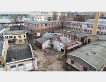 Luftaufnahme eines alten Fabrik- und Industrieareals mit mehreren Backstein- und Putzgebäuden, teils beschädigten Dächern und einem offenen Innenhof mit Betonflächen. Rechts ragt ein hoher Schornstein neben langen Werkhallen hervor, im Hintergrund stehen weitere größere Gewerbebauten.