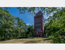 Backsteinturm mit rechteckigen Fenstern und spitzem Dach in einem bewaldeten Park. Der Turm ist von Bäumen und einer Wiesenfläche umgeben, die teilweise von Schatten bedeckt ist.
