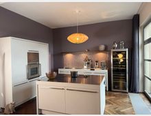 Moderne Küche mit einer weißen Kochinsel, eingebautem Ofen und einem Kühlgerät für Weinflaschen. Dunkelviolette Wände, ein Hängependel als Lampe, hochwertiger Holzfußboden und ein großes Fenster mit grauem Vorhang prägen den Raum.