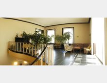 Heller Aufenthaltsbereich mit großen Fenstern, Zimmerpflanzen und Sitzmöbeln, darunter Sessel, ein Sofa und ein kleiner Tisch; balustradengeländer einer geschwungenen Treppe im Vordergrund.