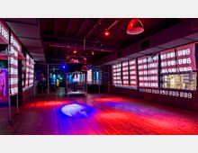 Ein Nachtclub- oder Veranstaltungsraum mit dunkler Holzbodenfläche, einer Spiegelkugel in der Mitte und Wänden, die mit leuchtenden Zahlenmustern beleuchtet sind. Überkopf befinden sich rote und blaue Lichtquellen, die den Raum in eine bunte Atmosphäre tauchen.