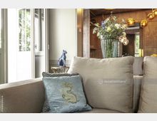 Modernes Wohnzimmer mit einem cremefarbenen Sofa, darauf ein verziertes hellblaues Kissen. Im Hintergrund stehen eine blauglasierte Katzenfigur auf einem kleinen Beistelltisch sowie eine Vase mit frischen Blumen auf einem Sideboard. Holzvertäfelung und goldfarbene Hängeleuchten definieren die Raumgestaltung.