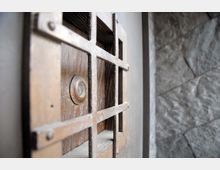 Nahaufnahme eines Metalgitters vor einem kleinen Sichtfenster in einer hölzernen Tür, daneben eine grob behauene Steinmauer.