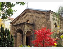 Backsteingebäude einer kleinen Kirche mit Rundbogenportal und Kreuz auf dem Dach. Im Vordergrund sind ein roter Laubbaum und weitere Pflanzen, die von grünen Baumzweigen umrahmt werden.