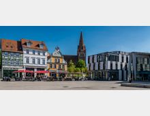 Stadtplatz mit historischen und modernen Gebäuden, darunter Fachwerk- und Barockfassaden auf der linken Seite sowie ein moderner Bau rechts. Im Hintergrund ist der Turm einer Kirche zu sehen, während am Platz Cafés mit Sonnenschirmen sowie parkende Fahrräder sichtbar sind.