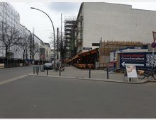 Straßenszene in einer Stadt mit einer Kreuzung und Gehwegen. Links sind Gebäude mit Baugerüsten und Planen bedeckt, rechts ein Café mit Sitzgelegenheiten im Freien und ein Container mit Werbung für kostenlose Corona-Schnelltests. Fahrräder und Schilder stehen entlang des Gehwegs.
