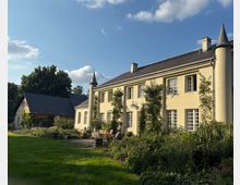 Zweistöckiges, helles Landhaus mit dunklem Satteldach und turmartigen Eckpfeilern mit Spitzdächern, davor eine Terrasse und ein bepflanzter Garten. Links steht ein Nebengebäude mit Schieferdach; die Anlage ist von Rasenflächen und Bäumen unter blauem Himmel umgeben.