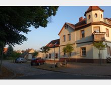 Wohnstraße mit einem zweistöckigen Gebäude im traditionellen Stil, gelber Putz mit roten Dachziegeln und einem kleinen Turm als architektonisches Detail. Vor dem Gebäude stehen mehrere junge Bäume, geparkte Autos und eine Holzbank auf einem gepflasterten Gehweg.